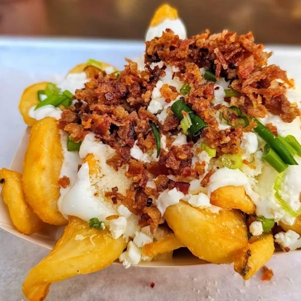 Close-up of golden potato wedges topped with white sauce, crumbled bacon, and chopped green onions.