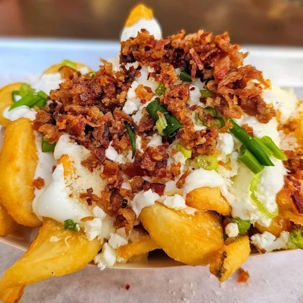 Close-up of golden fries topped with white sauce, crumbled bacon, cheese, and green onions.