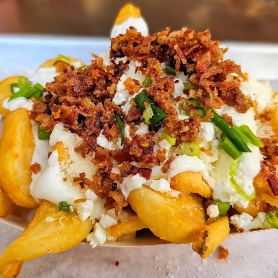 Close-up of golden fries topped with white sauce, crumbled bacon, cheese, and green onions.